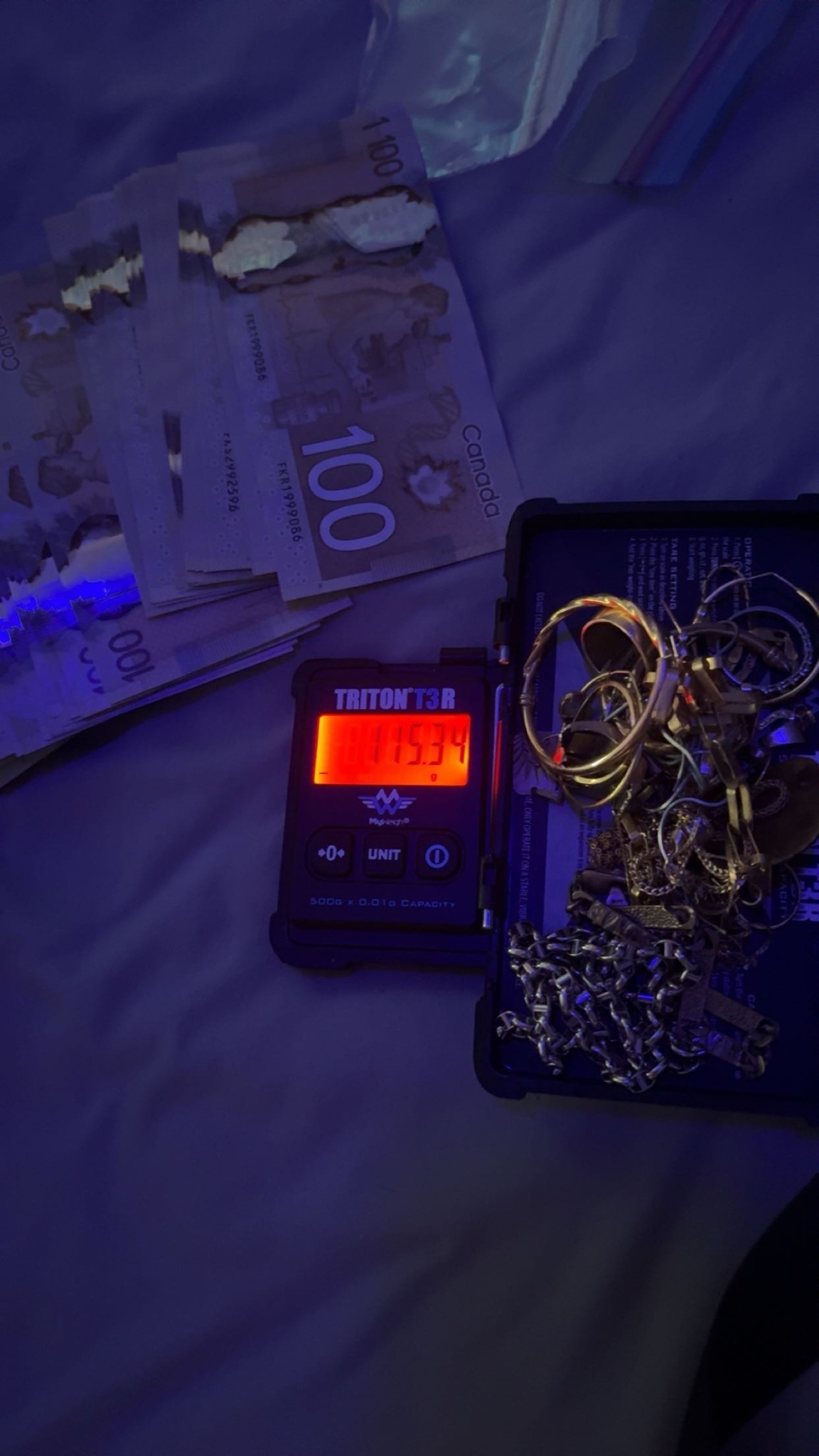 Gold jewellery on a red digital scale beside Canadian banknotes under purple light