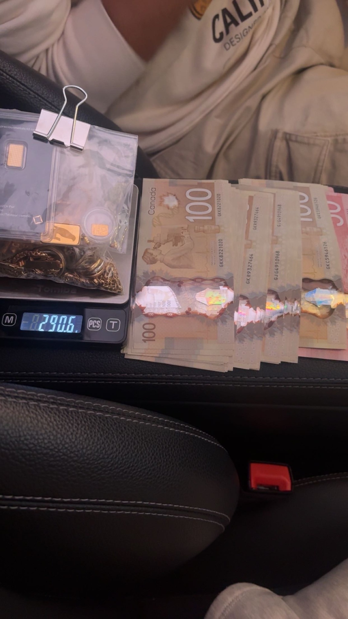 Gold bars and jewellery on a digital scale beside Canadian banknotes inside a vehicle